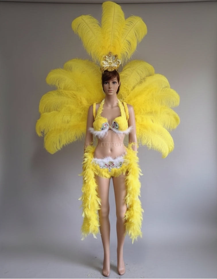 A woman modeling a bright yellow carnival costume with a large feathered headdress, matching feathered shoulder pieces, a feathered belt, and a small gold crown, standing against a plain indoor wall.