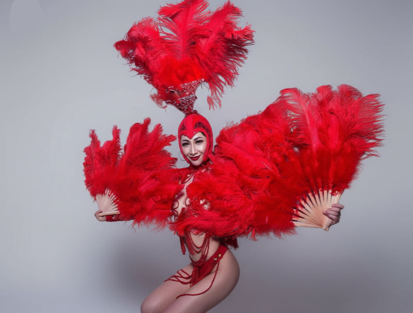 A performer in a striking red feathered costume with matching headpiece and large feathered fan-like accessories, posed on a dark stage under blue and red lighting.