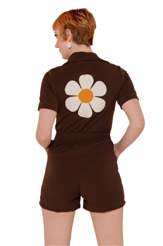A person wearing a brown romper with a large white and yellow flower applique on the back, short sleeves, and front hand pockets.