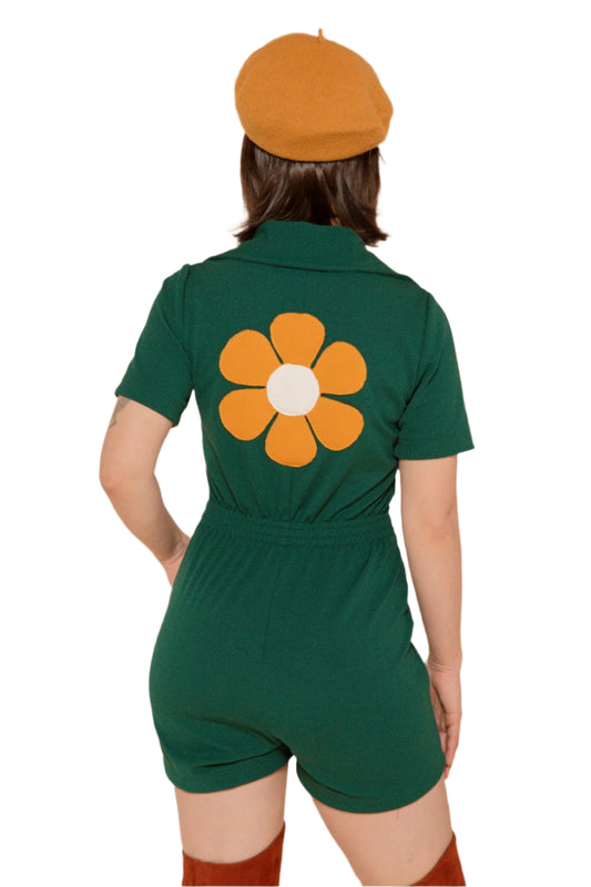 A person wearing a green romper with a hand-cut flower applique on the back, short sleeves, and an elasticized waist. They are also wearing a beret and brown shoes.